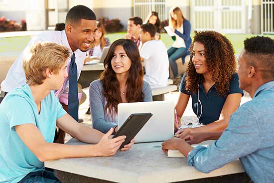 Teacher interacts with group of high school students using various digital devices.